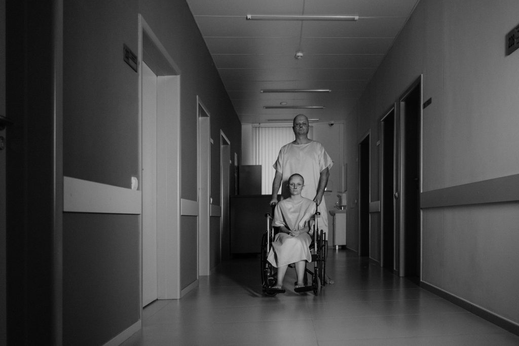 Two patients in hospital gowns, a man, and a wheelchair user, in a hospital hallway.
